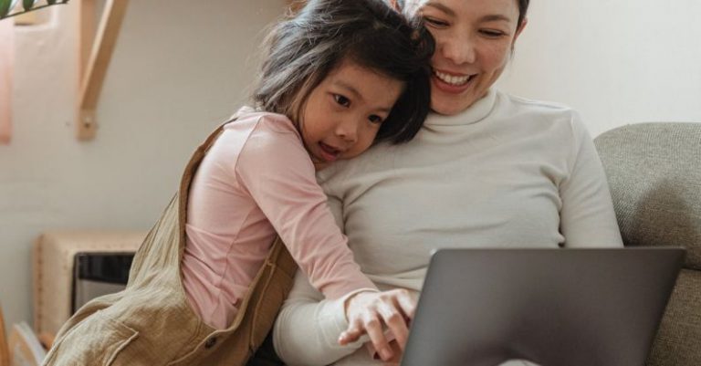 User-Generated Content Policies - Cheerful smiling Asian woman browsing modern netbook while hugging with cute content daughter on comfy sofa in cozy living room