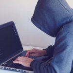 Cyber Threats - Side view of unrecognizable hacker in hoodie sitting at white table and working remotely on netbook in light room near wall