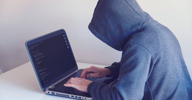 Cyber Threats - Side view of unrecognizable hacker in hoodie sitting at white table and working remotely on netbook in light room near wall